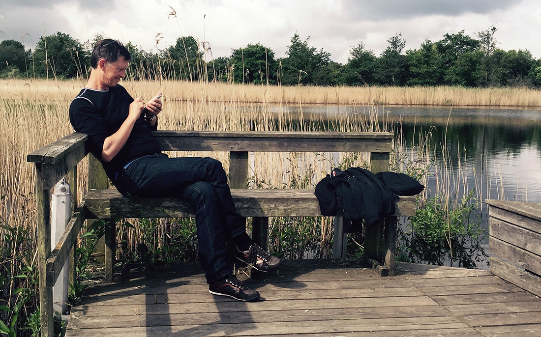 Mandlig besøgende på bænk ved sø læser om stedet på sin mobil